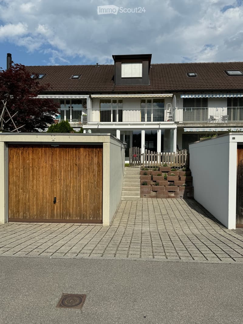 Two-story house with a tiled roof, large windows, and a balcony. Features a wooden garage door and a paved driveway.