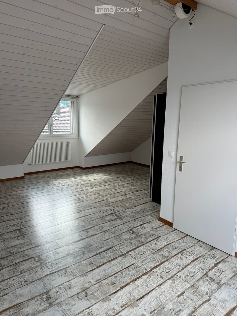 Empty attic room with wooden floor and slanted wooden ceiling, white walls, and a window letting in natural light.