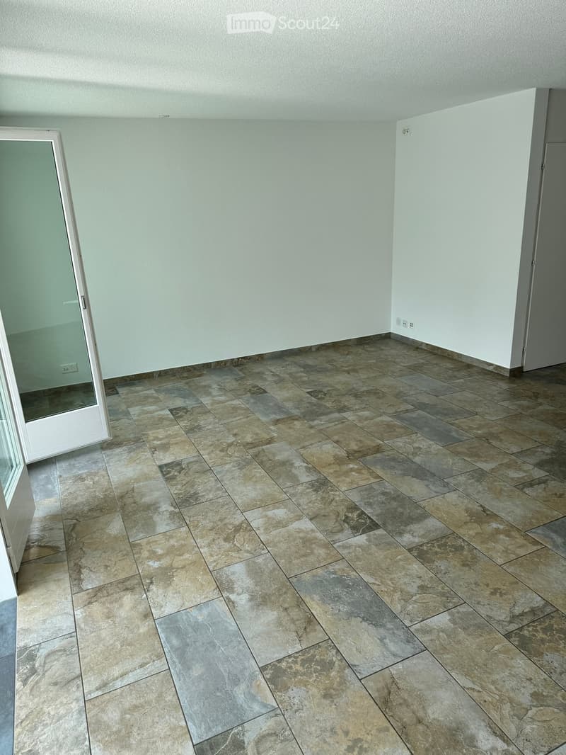 Empty room with light-colored walls and a large, patterned tile floor. The room features a white door frame and a glass door leading to another space.