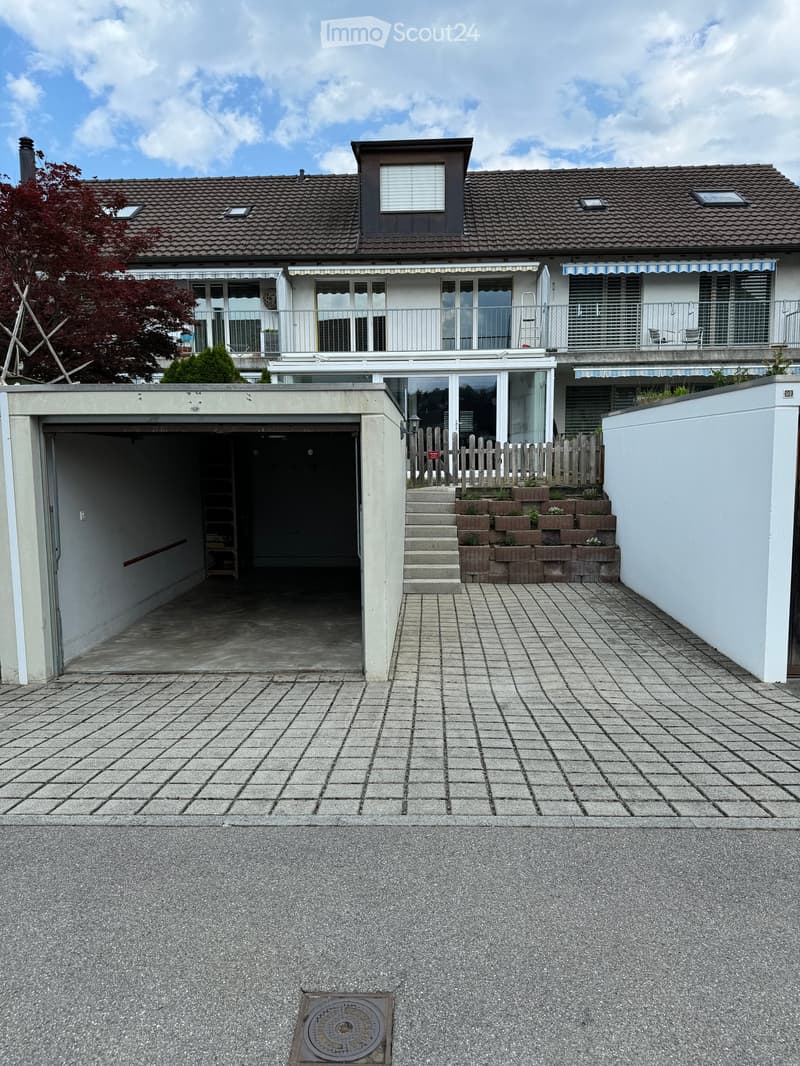 Two-story house with a garage, raised ground floor, and balcony. The house features a tiled roof, large windows, and a small garden area with raised planters.
