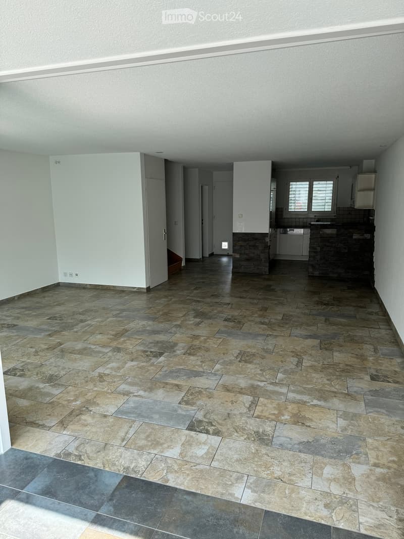 Empty room with beige tiled floor, white walls, and visible kitchen area with cabinets and appliances in the background.