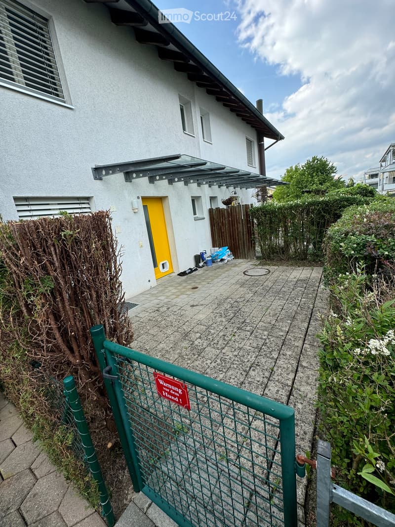 Two-story house with white exterior, yellow front door, and a covered entrance area. Green metal gate with warning sign, surrounded by bushes and a paved pathway.