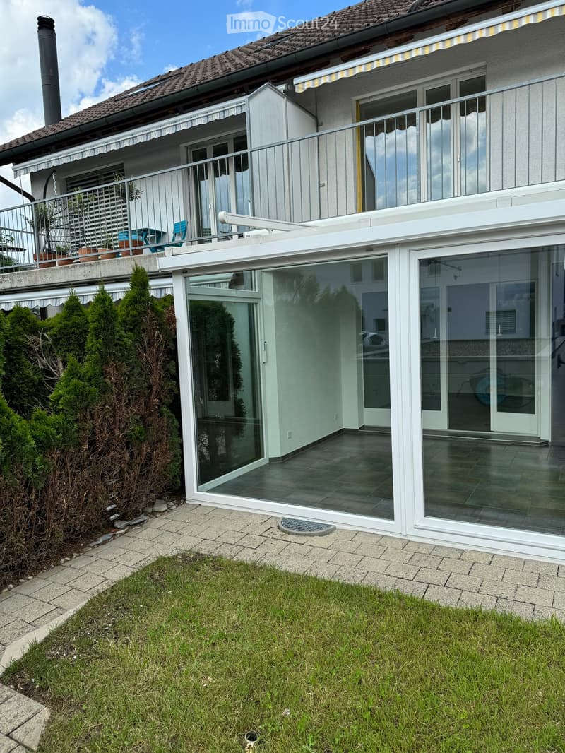 Garden area with a well-maintained lawn, a paved pathway, and a modern house with large glass doors and a balcony. The house features a tiled roof and a small patio area with outdoor furniture.