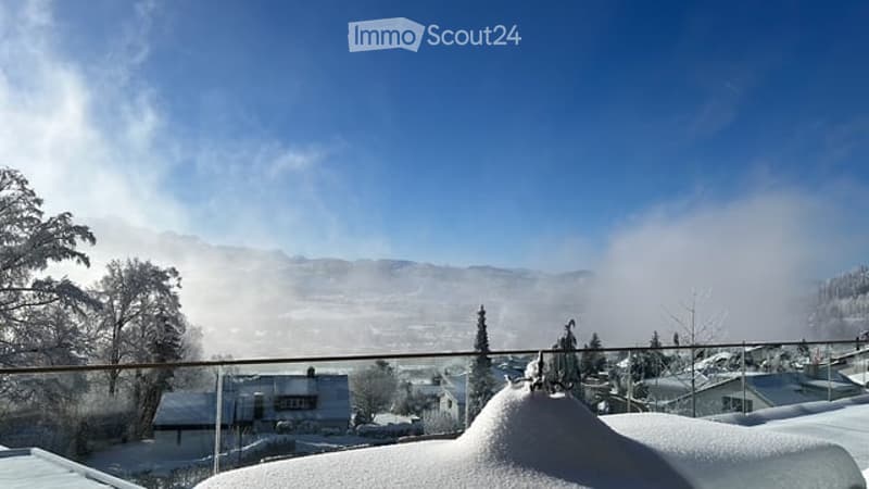 A snowy balcony or terrace overlooking a mountain landscape with a town below, clear blue sky, and snow-covered roofs.