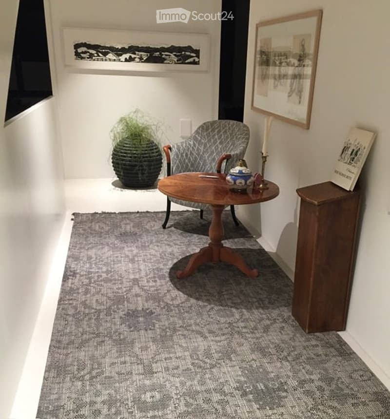 A small, well-lit room with a grey armchair, a wooden coffee table, a grey rug, a decorative plant, and a wooden cabinet with a book on top.