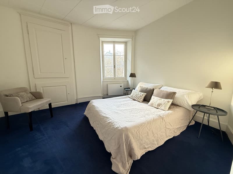 Cozy bedroom with a large bed, nightstands, a chair, and table lamps. The room features blue carpeting and white walls.