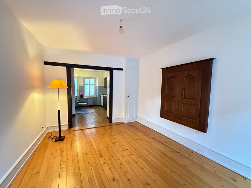 Empty room with wooden flooring, white walls, and a large wooden cabinet on one wall. A floor lamp is placed near the doorway, and an open doorway leads to another room with visible furniture.