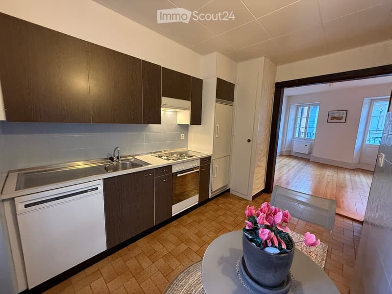 Modern kitchen with dark wooden cabinets, stainless steel appliances, and a small round table with a flower pot in the foreground. The adjacent room has wooden flooring and large windows.