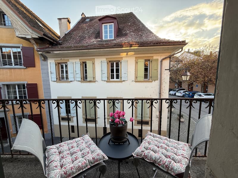 Balcony with two chairs, a small round table, and a flower pot with pink flowers. The view shows a charming European street with traditional houses and parked cars.