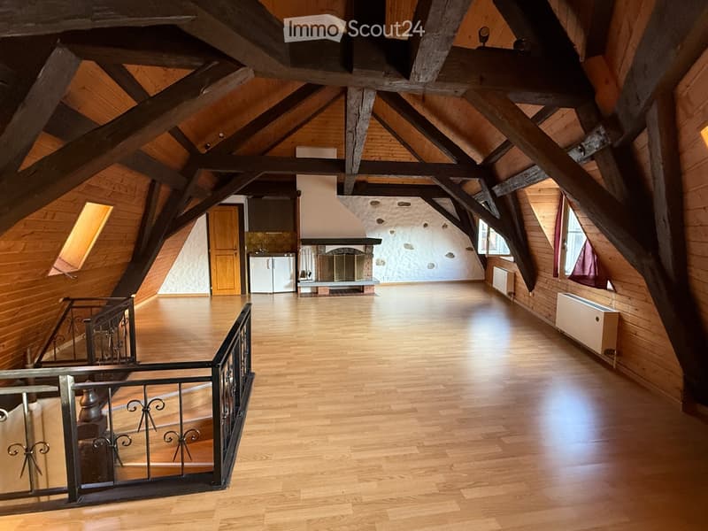 Spacious attic room with wooden beams, polished wooden floor, fireplace, refrigerator, and metal railing leading to another level.
