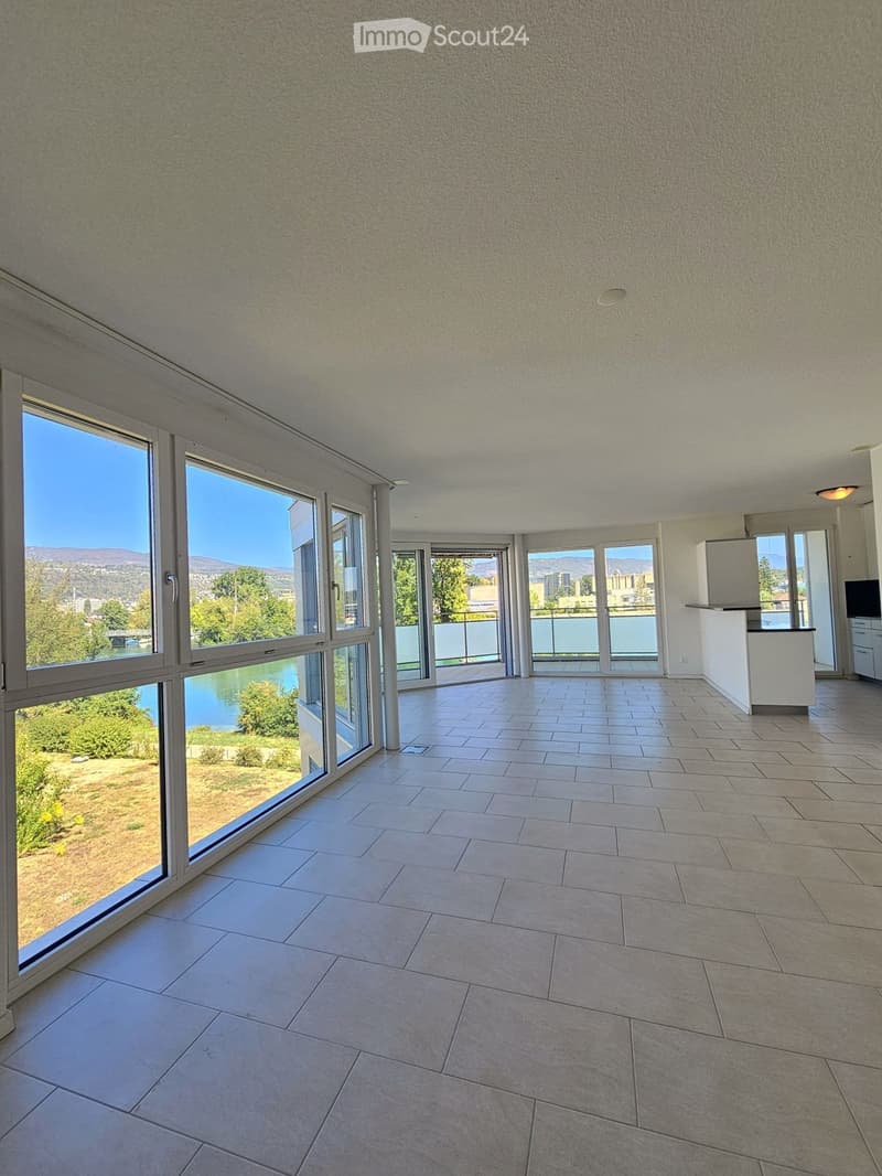 Empty room with large windows offering a scenic view of a lake and surrounding landscape. The floor is tiled, and the walls are white.