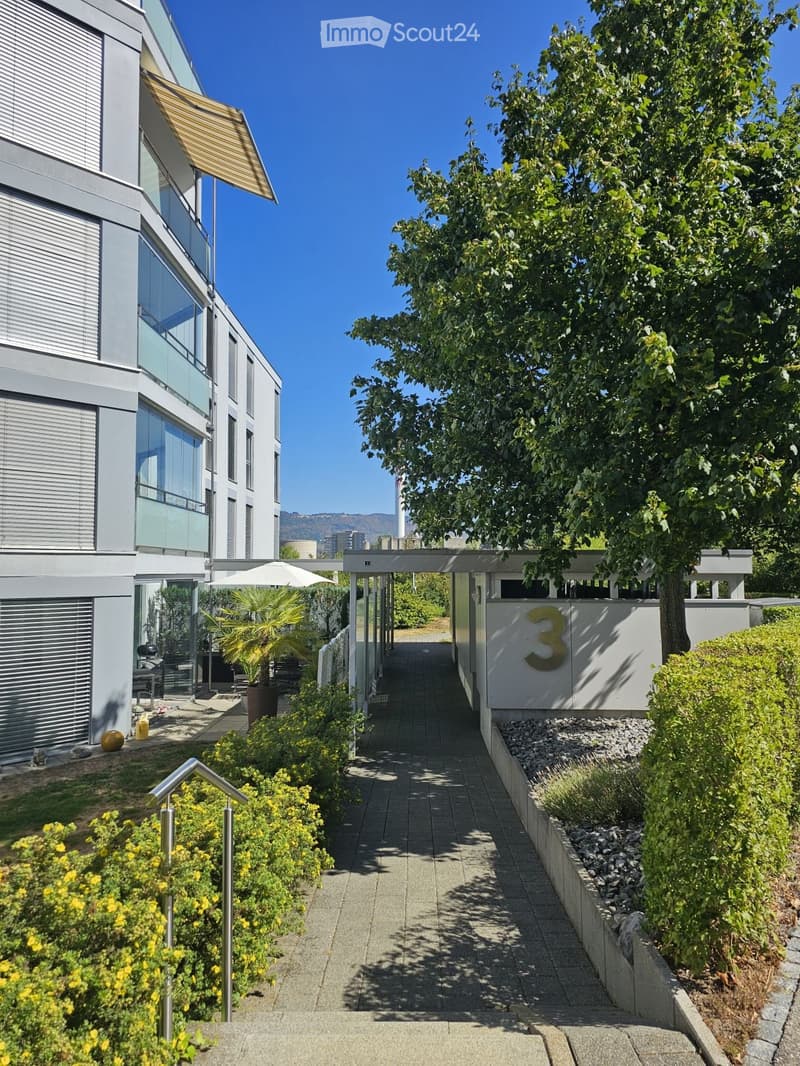 A modern residential complex with a paved pathway leading to a building entrance, surrounded by greenery and a large tree. The building features large windows and a sleek design.