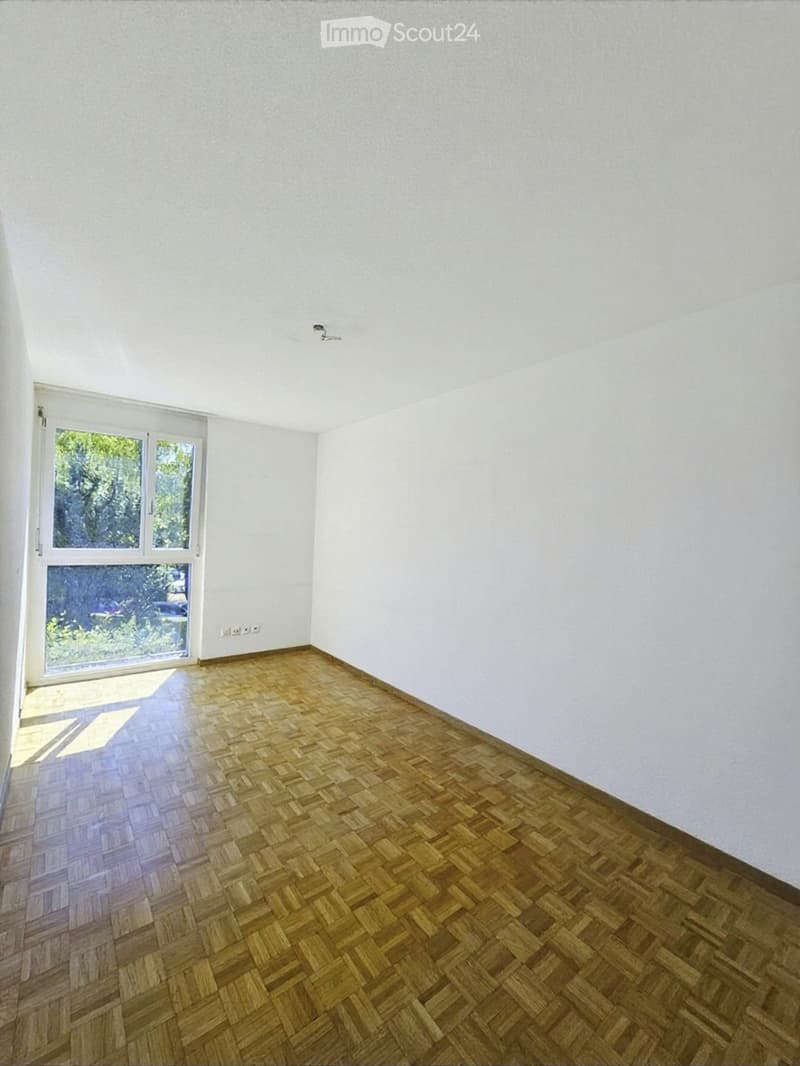 Empty room with white walls, wooden herringbone flooring, and large window with white frame. No furniture or equipment present.