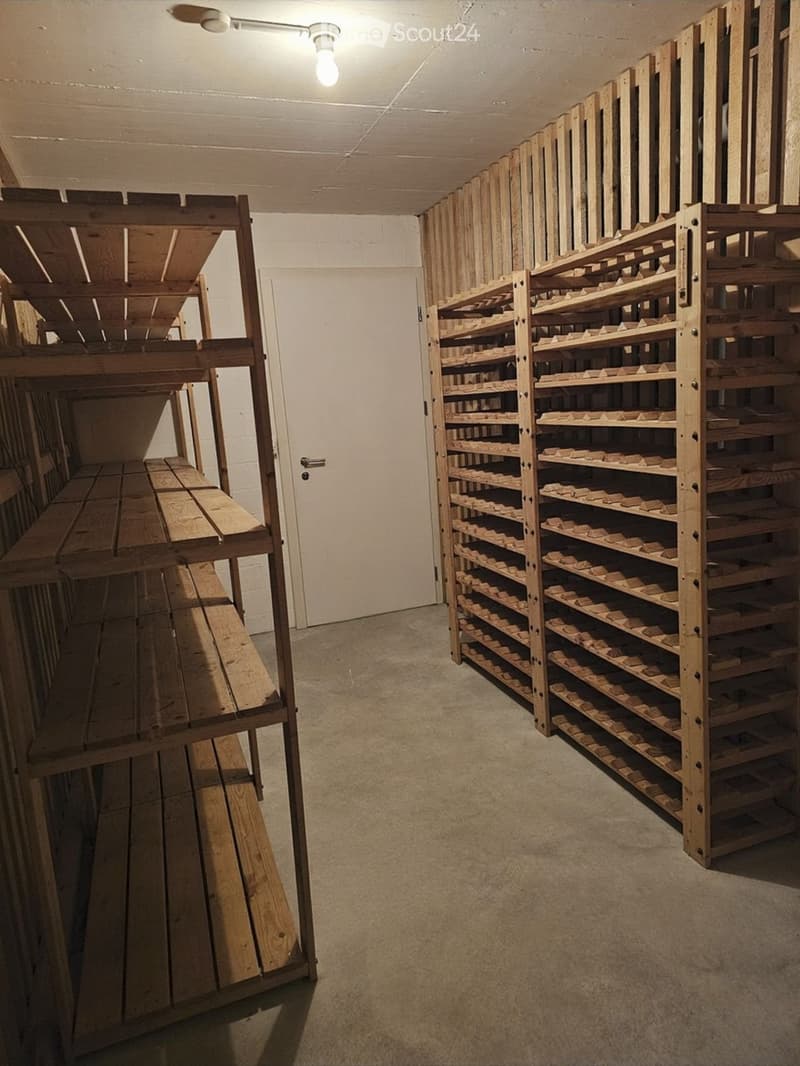 Empty storage room with wooden shelving units and a door, concrete floor, and exposed ceiling beams.