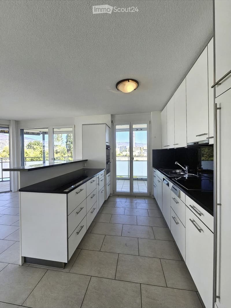 Modern kitchen with white cabinets, black countertops, and a central island. Features a sink, faucet, and oven, with large windows and a sliding glass door leading to a balcony.