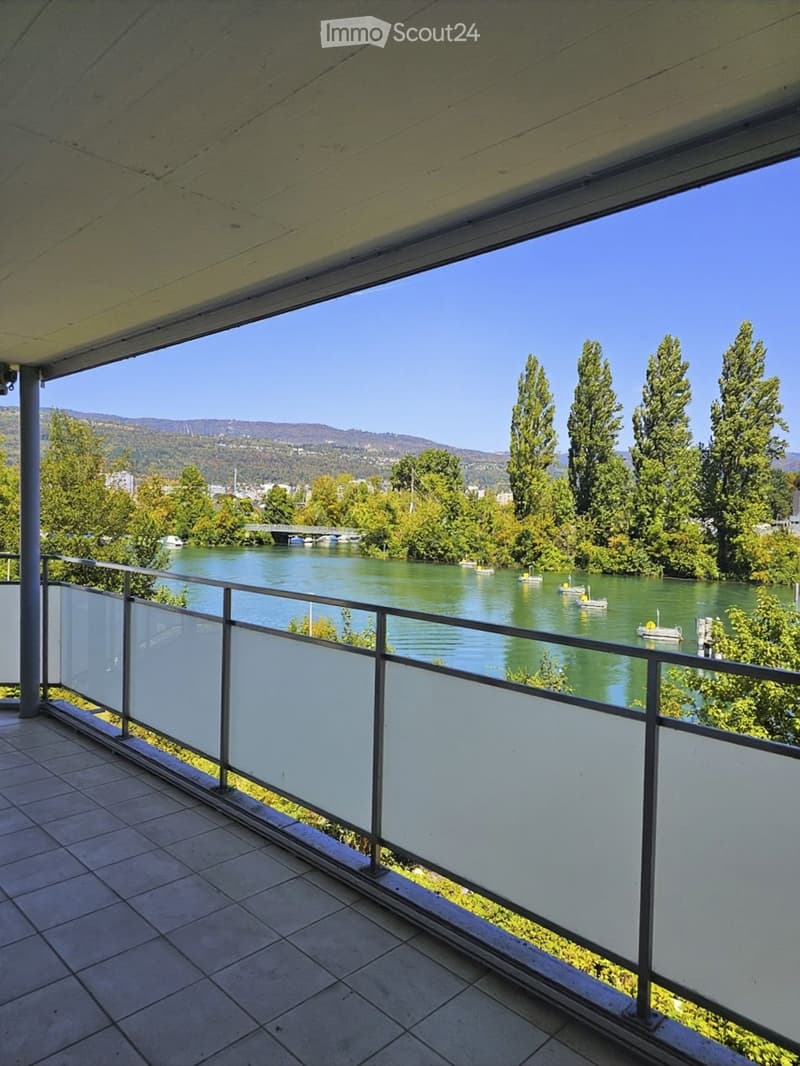 Balcony with a modern glass railing, overlooking a river with boats and surrounding greenery and hills.