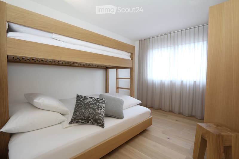 A modern, minimalist bedroom with a wooden bunk bed, white bedding, and a decorative pillow. The room features light wooden flooring and sheer curtains on the windows.
