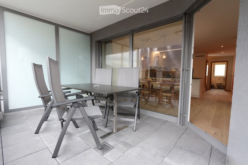 Modern balcony with tiled floor, large glass sliding doors, and a dining setup including a table and chairs.