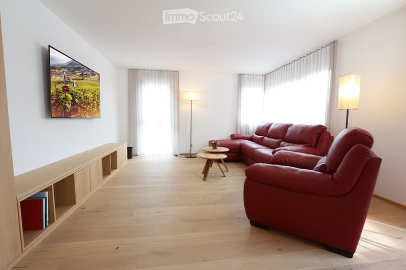 Modern living room with a red leather sofa, armchair, coffee table, floor lamps, and a TV mounted on the wall. The room features large windows with white curtains and a wooden shelf with books.