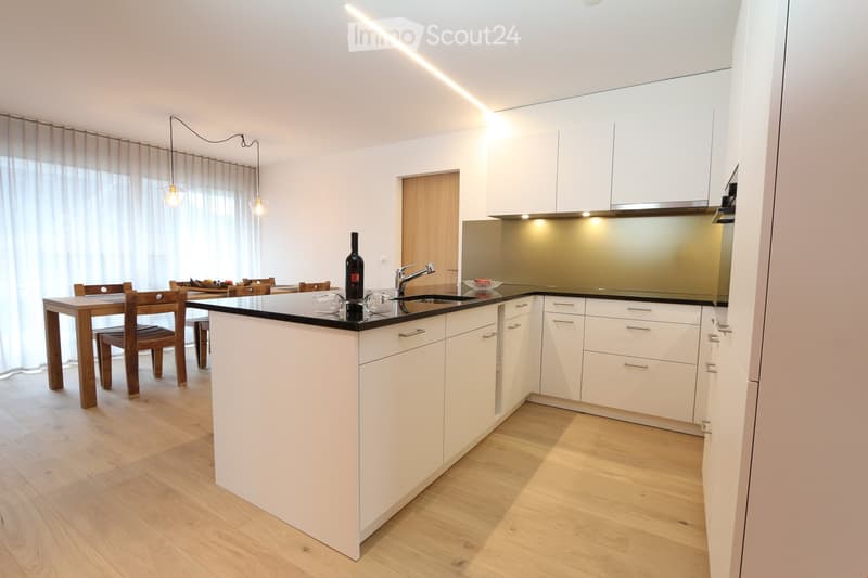 Modern kitchen with white cabinets, a central island, and a dining area with wooden chairs and a table. The kitchen features a sleek black countertop, a sink with a modern faucet, and pendant lights hanging from the ceiling.
