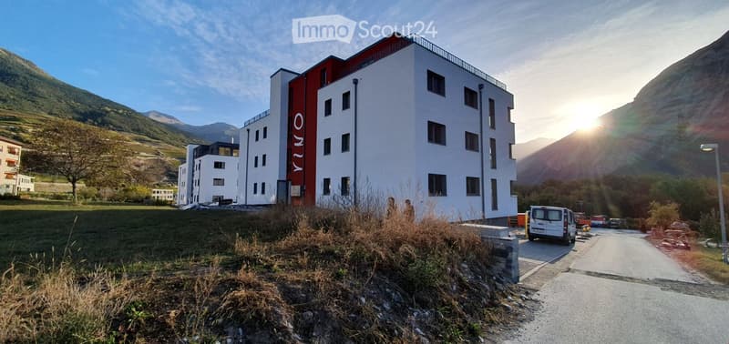 Modern multi-story building with white and red facade, located in a scenic mountainous area with a parking lot and surrounding greenery.