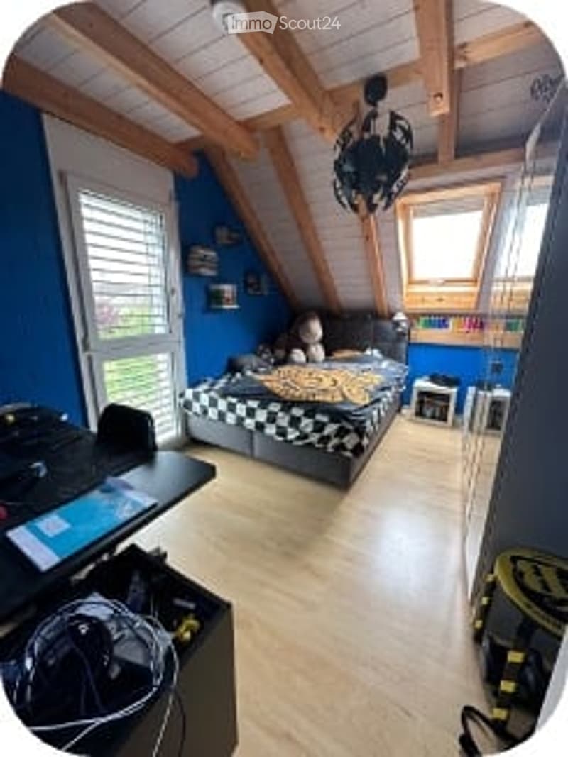 Cozy attic bedroom with wooden beams, blue walls, a bed with a checkered duvet, a desk with a chair, and a bookshelf.