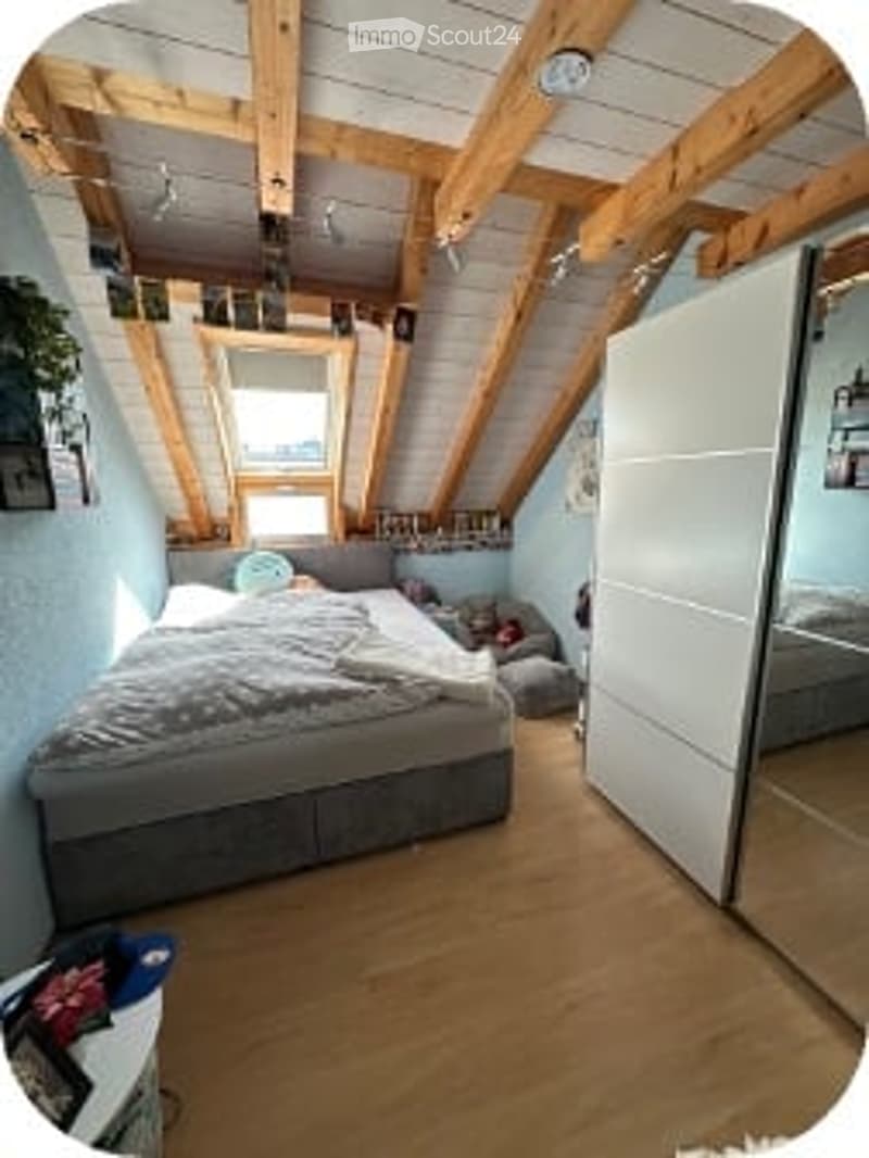 Slanted wooden ceiling beams, light blue walls, grey bed with white bedding, white wardrobe, nightstand, and potted plant on the floor.