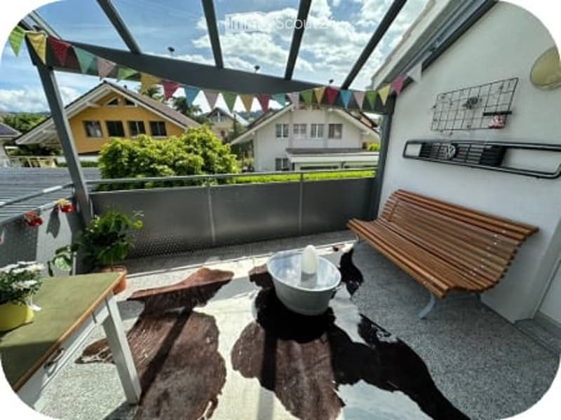 A modern rooftop terrace with a wooden bench, small table, and decorative elements, overlooking a residential area with houses and greenery.