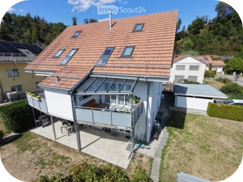 A modern two-story house with a red-tiled roof, white exterior, and a large balcony terrace on the first floor. The house features multiple windows and a small garden area.