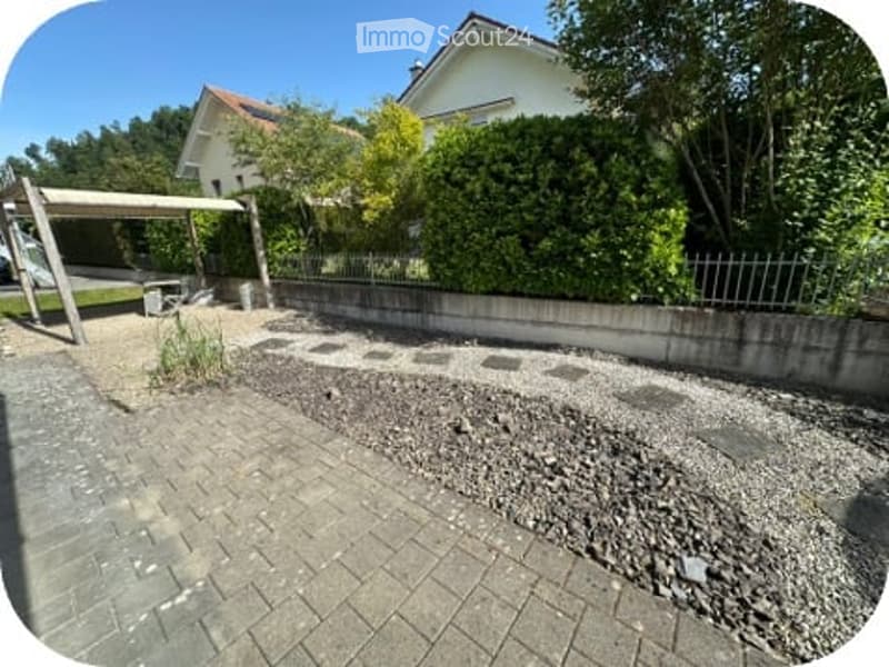 Garden area with gravel, patio, small wooden shelter, and a white house in the background.