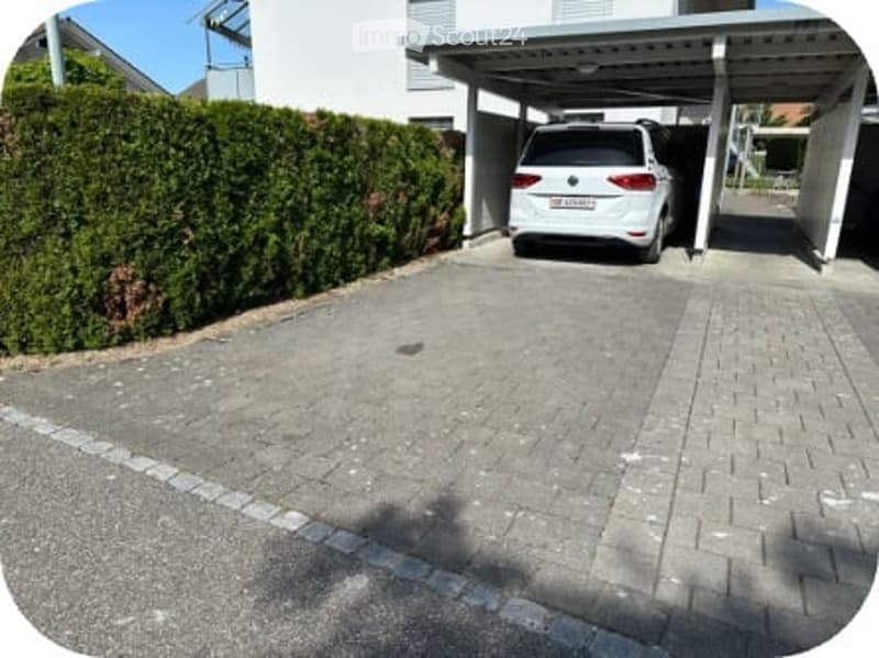 Driveway with a parked car under a covered garage area, surrounded by a neatly trimmed hedge and a modern building in the background.