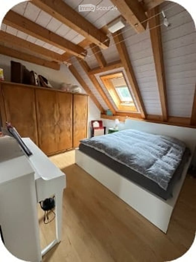 Cozy attic bedroom with wooden beams, skylight window, white bed, wardrobe, and desk.