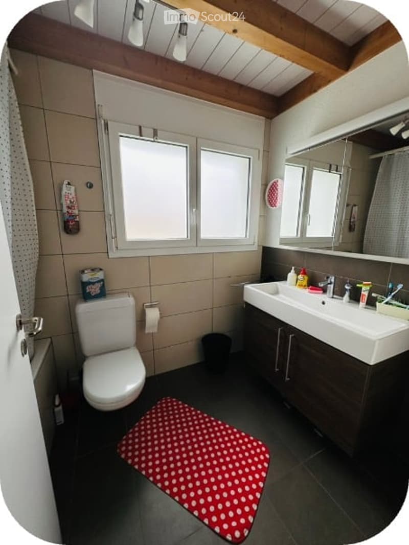 Bathroom with a toilet, sink, mirror, and a red and white polka dot bathroom mat on the floor. The room features wooden beams on the ceiling and tiled walls.