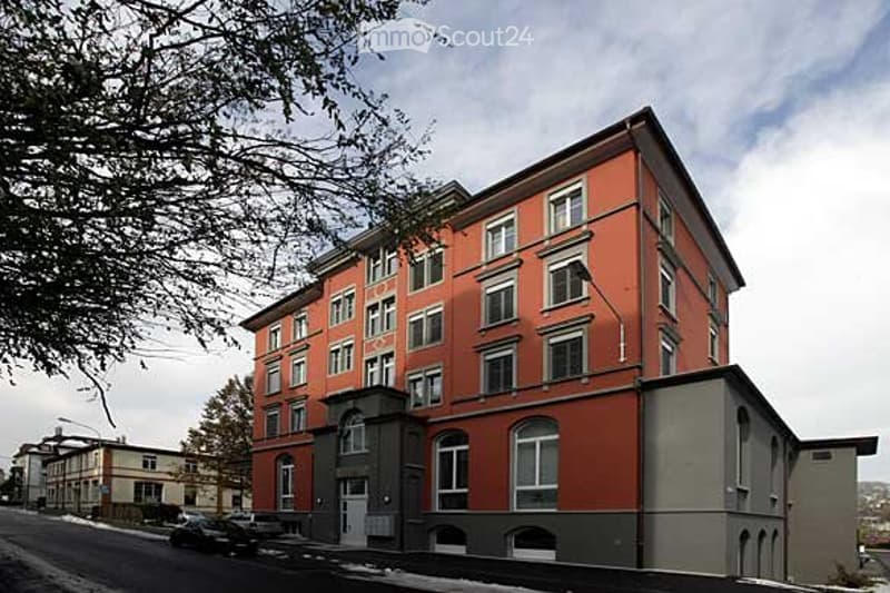 Multi-story building with red brick facade and white-framed windows, featuring a central entrance and adjacent parking area.