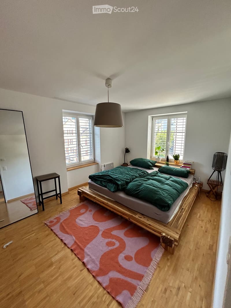 Modern bedroom with wooden floor, large bed with green duvet, colorful rug, mirror, small stool, table lamp, potted plants, and books on nightstand.