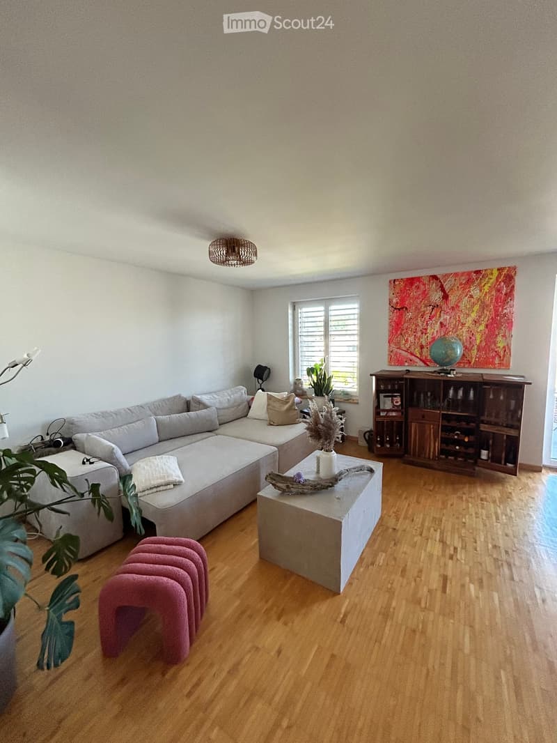 Modern living room with light-colored sofa, coffee table, pink stools, bookshelf, and decorative items.