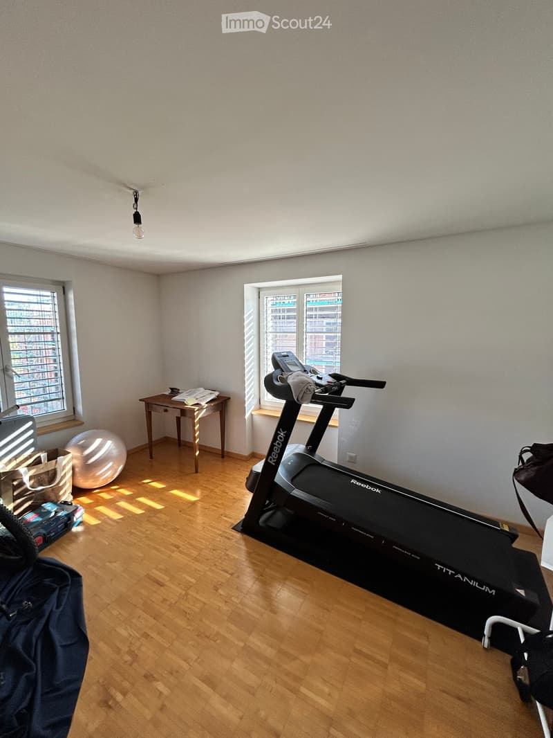 A room with wooden flooring, a treadmill, a large exercise ball, a small table, and a handbag. The room has two windows with blinds and a hanging light fixture.