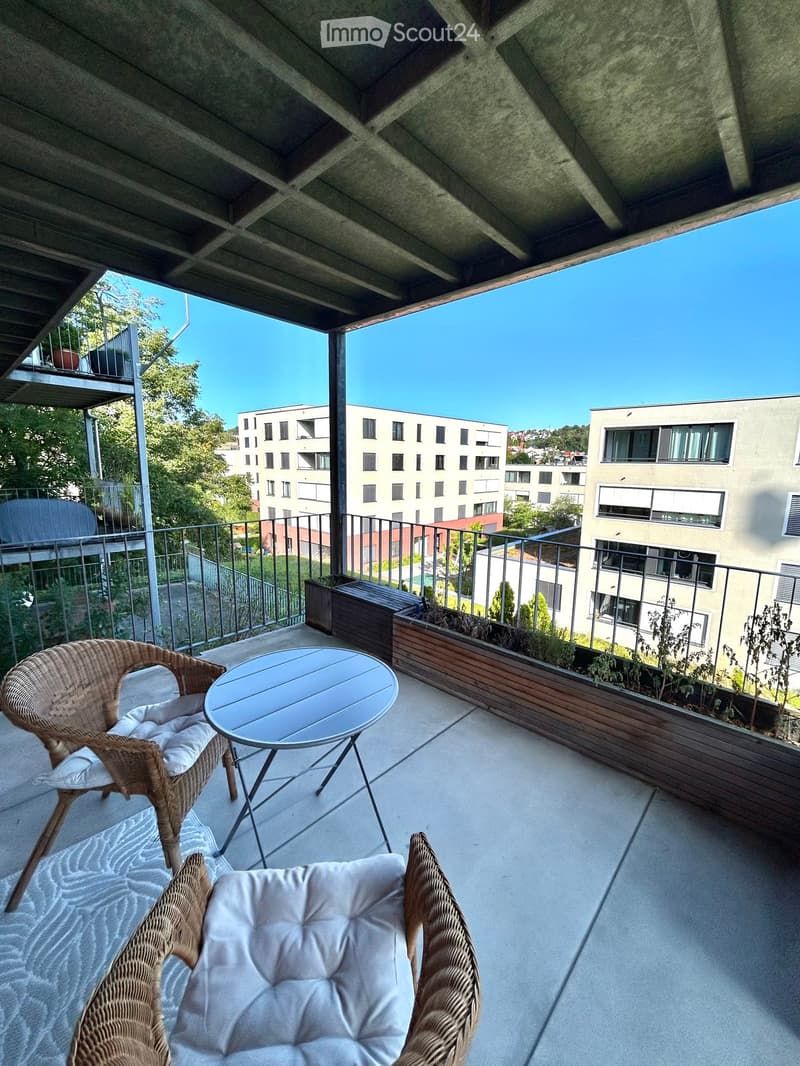 Balcony with a round table, two wicker chairs, and a view of modern buildings and greenery.