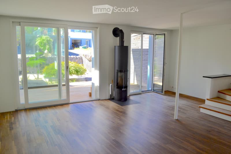 Modern living room with wooden flooring, large glass doors leading to a patio, and a central black wood-burning stove.