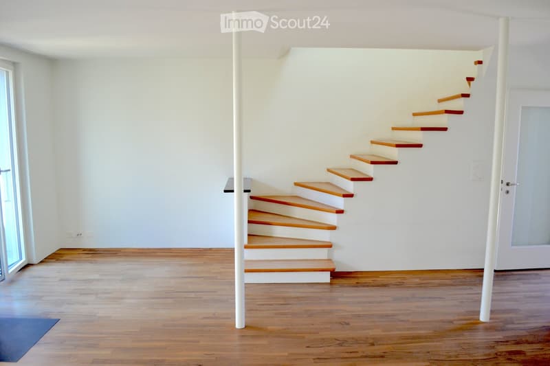 Empty room with wooden flooring, white walls, and a staircase leading upwards. The room has large windows and a door on the right side.