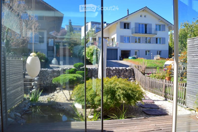 View from inside a house looking out onto a well-maintained garden with a small pond, stone pathways, shrubs, and a wooden fence. A white house with shutters and a garage is visible in the background.