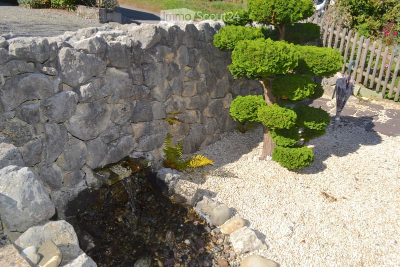 A small garden area with a stone wall, a small water feature, neatly trimmed shrubs, and a gravel pathway.