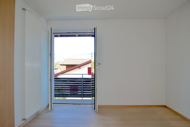 Empty room with light-colored walls and wooden flooring, featuring an open sliding door leading to a balcony with a metal railing and a view of a neighboring house.