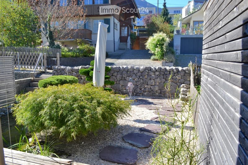 A well-maintained garden with a stone pathway, small water feature, shrubs, and a stone wall. The area is bordered by wooden fences and includes a covered patio umbrella and a small umbrella stand.