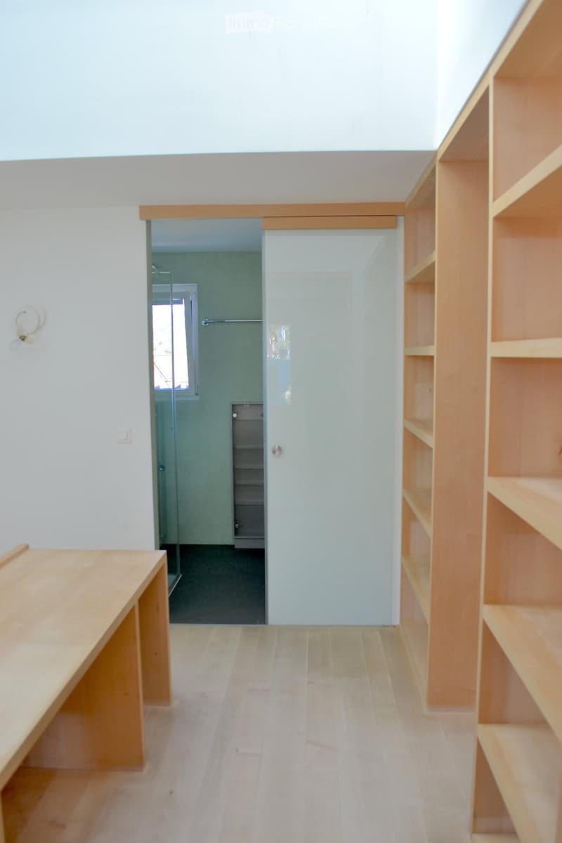 Empty room with light wooden flooring, white walls, built-in wooden shelving unit, and a sliding glass door leading to a small bathroom area with a shower and window.