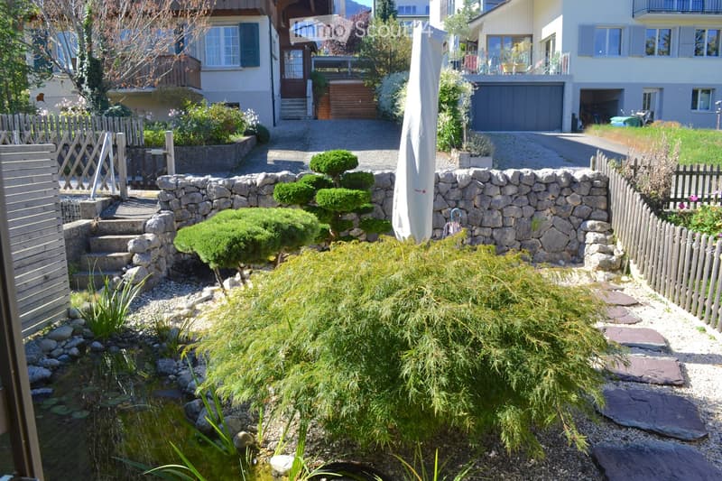 Well-maintained garden with a small pond, stone pathways, trimmed shrubs, and a stone wall. The garden features a variety of plants, a wooden fence, and a covered patio area in the background.