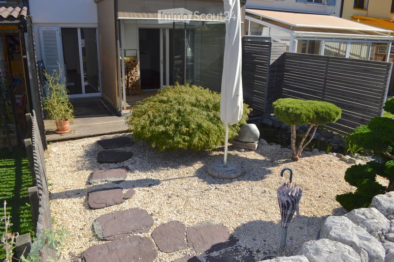 Garden with gravel, stepping stones, small trees, shrubs, a large umbrella, and a wooden deck connected to a house.