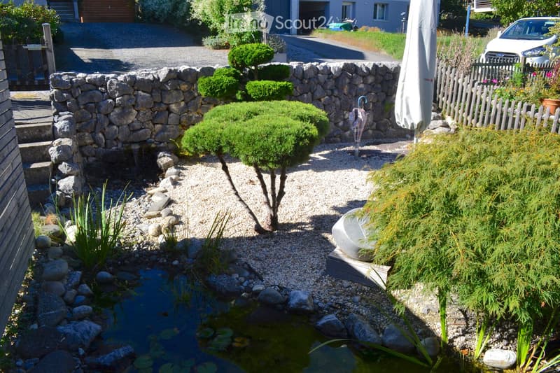 A well-maintained garden with a small pond, surrounded by rocks, gravel, and various plants, including a small tree and shrubs. A stone wall and wooden fence border the garden, with a closed umbrella and a car visible in the background.