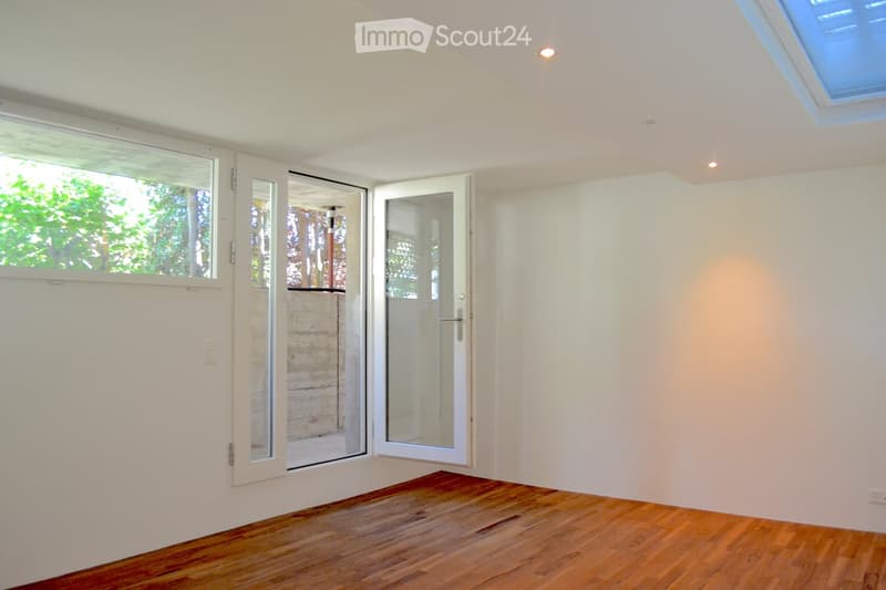 Empty room with wooden flooring, white walls, large windows, and a sliding glass door leading to an outdoor area.