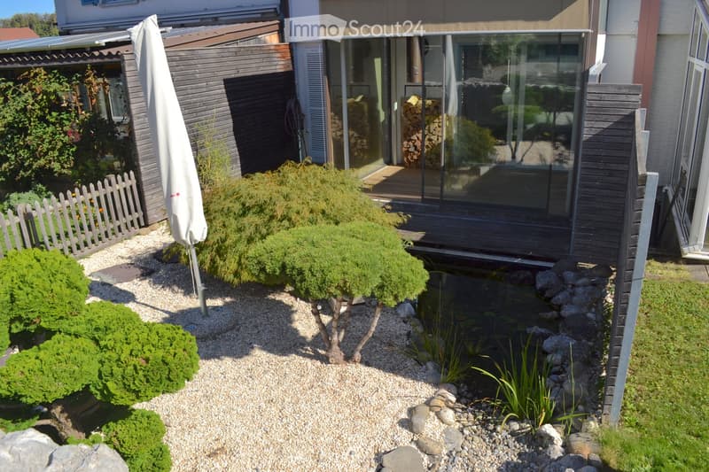 Well-maintained garden with gravel pathways, small pond, neatly trimmed shrubs, and a modern house with large glass windows and a covered patio area.